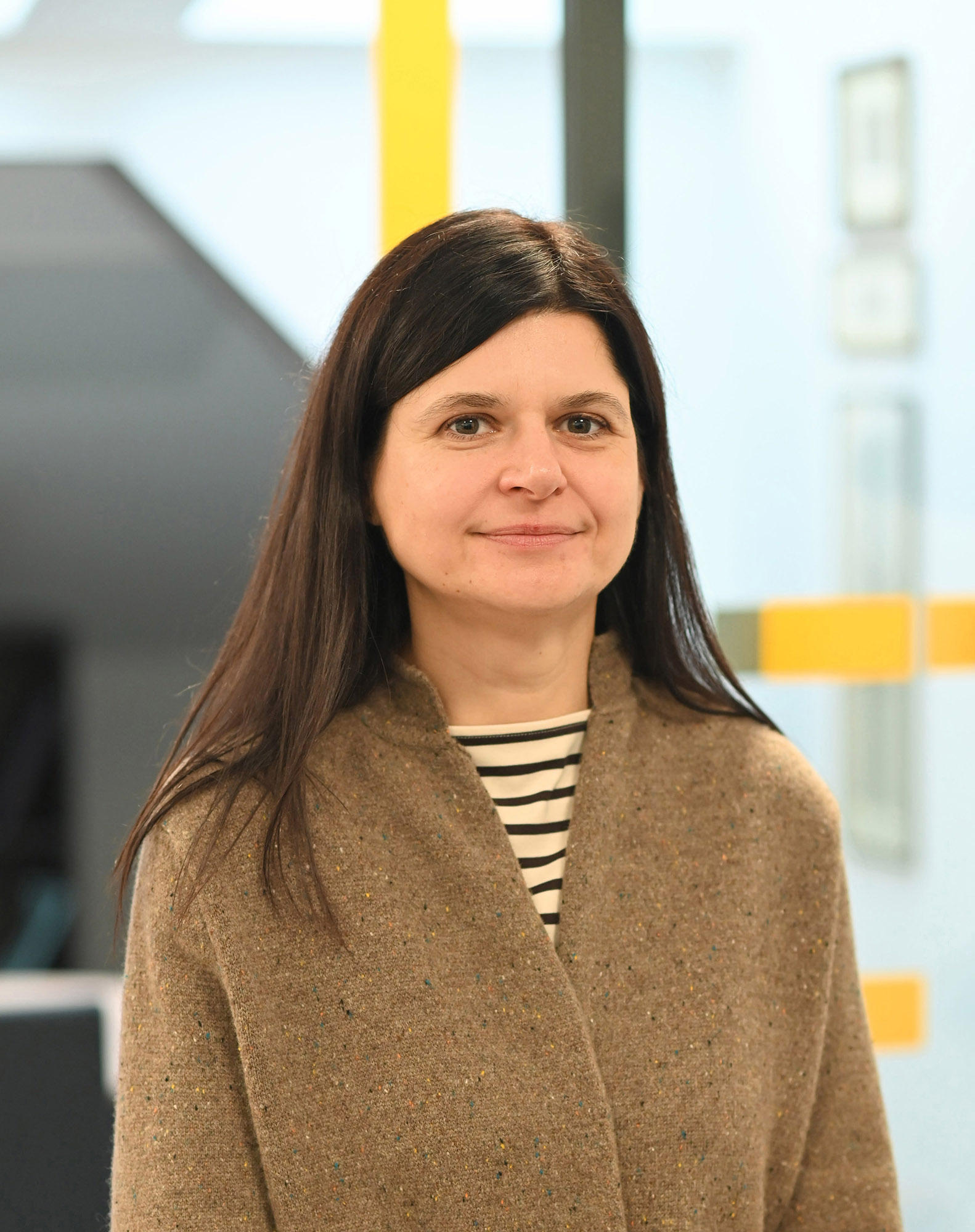 A middle-aged woman with long dark hair, wearing a brownish wool jacket with a high collar and a striped t-shirt. She is looking at the camera and smiling softly.