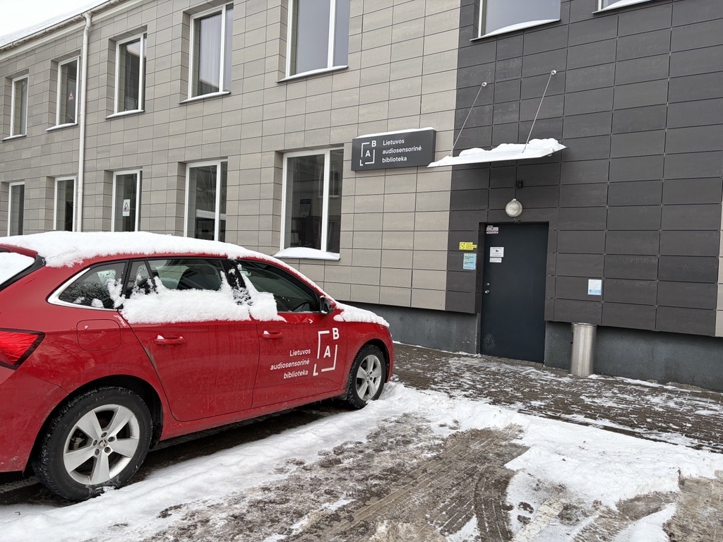 Įėjimo į LAB padalinį Panevėžyje nuotrauka. Žiema, dviaukštis pastatas, prie įėjimo stovi raudonas automobilis su LAB logotipu ant priekinių durelių.